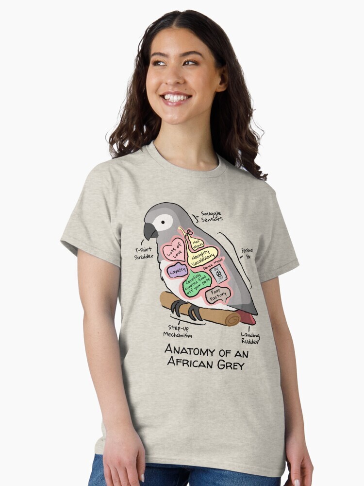 A brunette woman wearing a t-shirt  featuring a cartoon illustration titled “Anatomy of an African Grey” showing a cute African Grey  parrot with funny labeled anatomy sections.