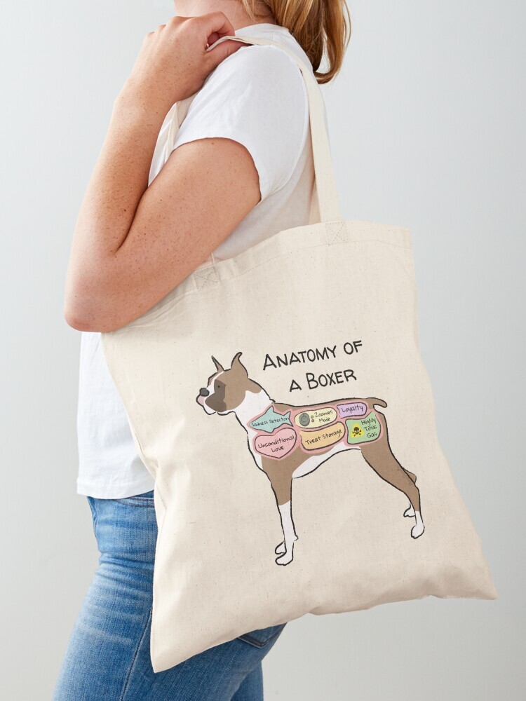 A woman wearing a white short sleeve t-shirt with a cotton tote bag on her shoulder. The tote bag features a cartoon illustration titled “Anatomy of a Boxer” showing a cute Boxer dog with funny labeled anatomy sections.