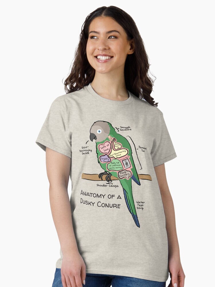 A brunette woman wearing a t-shirt  featuring a cartoon illustration titled “Anatomy of a Dusky Conure” showing a cute Dusky Conure bird with funny labeled anatomy sections.