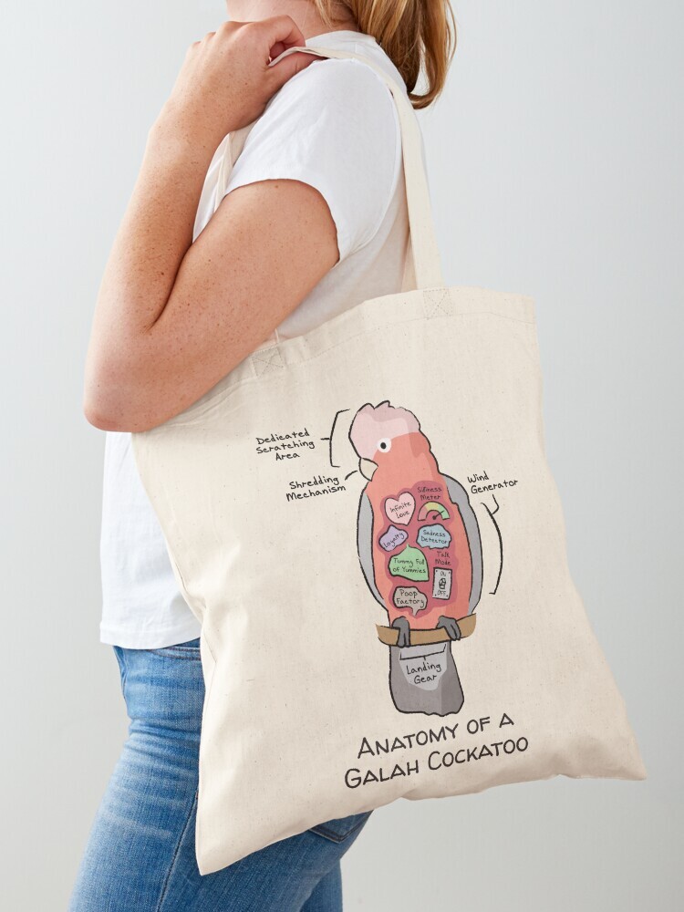 A woman wearing a white short sleeve t-shirt with a cotton tote bag on her shoulder. The tote bag features a cartoon illustration titled “Anatomy of a Galah Cockatoo” showing a cute Sun Conure bird with funny labeled anatomy sections.
