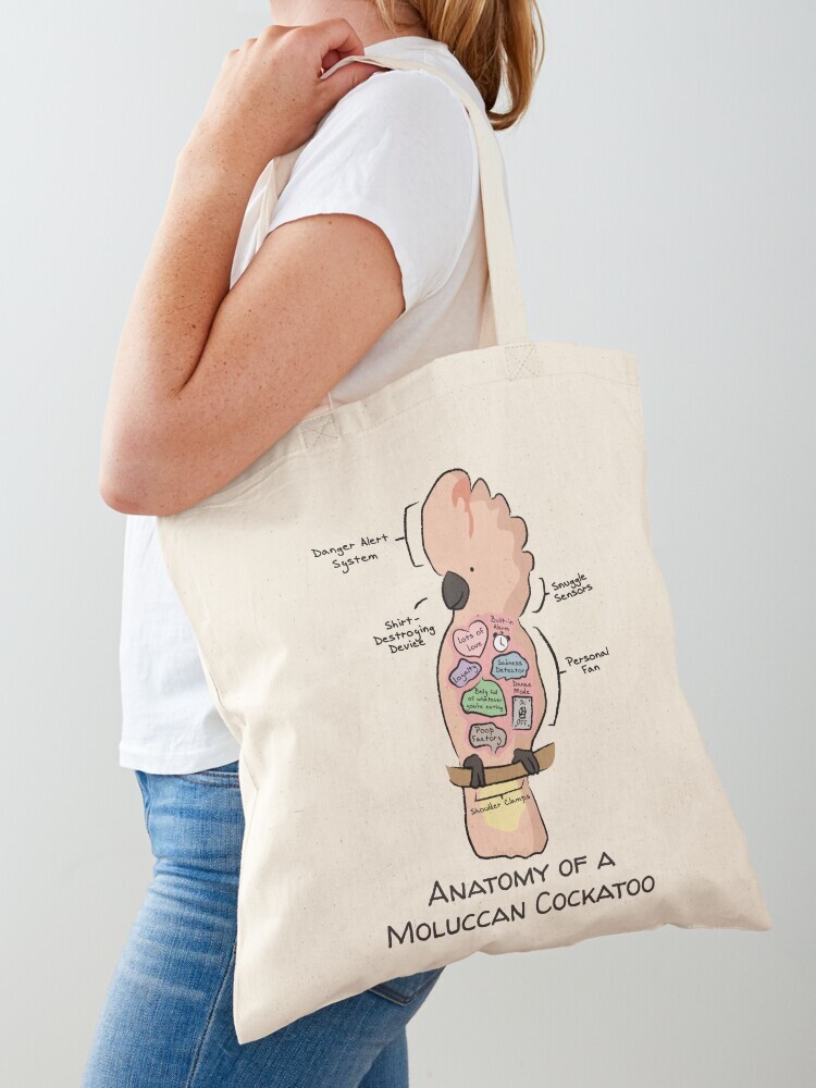 A woman wearing a white short sleeve t-shirt with a cotton tote bag on her shoulder. The tote bag features a cartoon illustration titled “Anatomy of a Moluccan Cockatoo” showing a cute Moluccan Cockatoo bird with funny labeled anatomy sections.