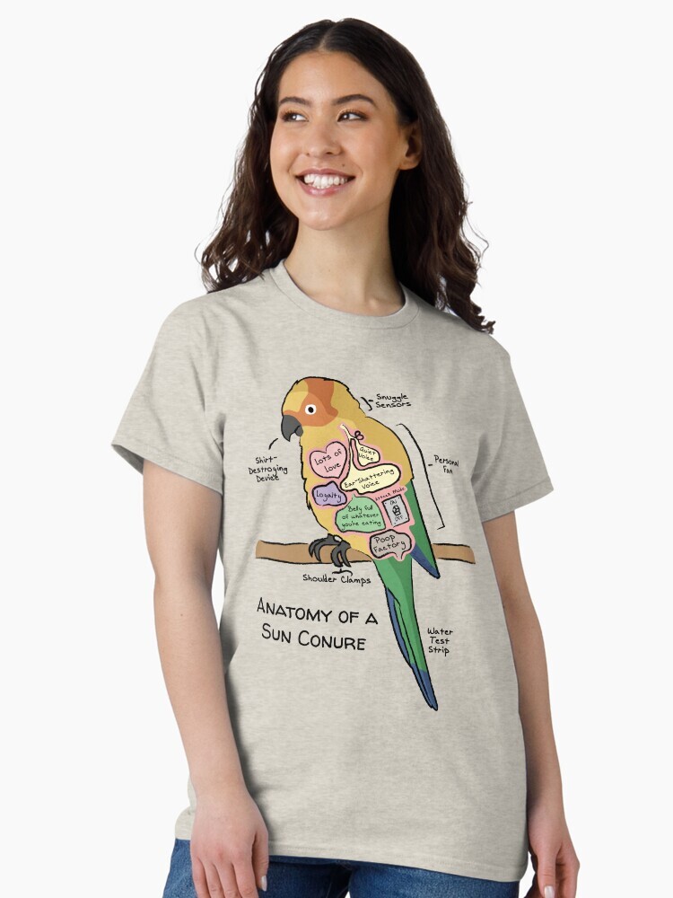 A brunette woman wearing a t-shirt  featuring a cartoon illustration titled “Anatomy of a Sun Conure” showing a cute Sun Conure bird with funny labeled anatomy sections.