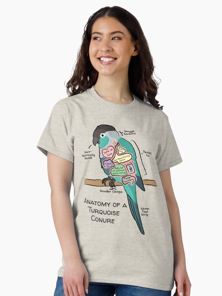 A brunette woman wearing a t-shirt  featuring a cartoon illustration titled “Anatomy of a Turquoise Conure” showing a cute Turquoise Conure bird with funny labeled anatomy sections.