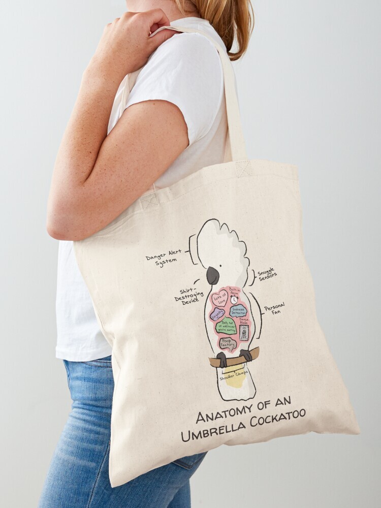 A woman wearing a white short sleeve t-shirt with a cotton tote bag on her shoulder. The tote bag features a cartoon illustration titled “Anatomy of a Sun Conure” showing a cute Sun Conure bird with funny labeled anatomy sections.