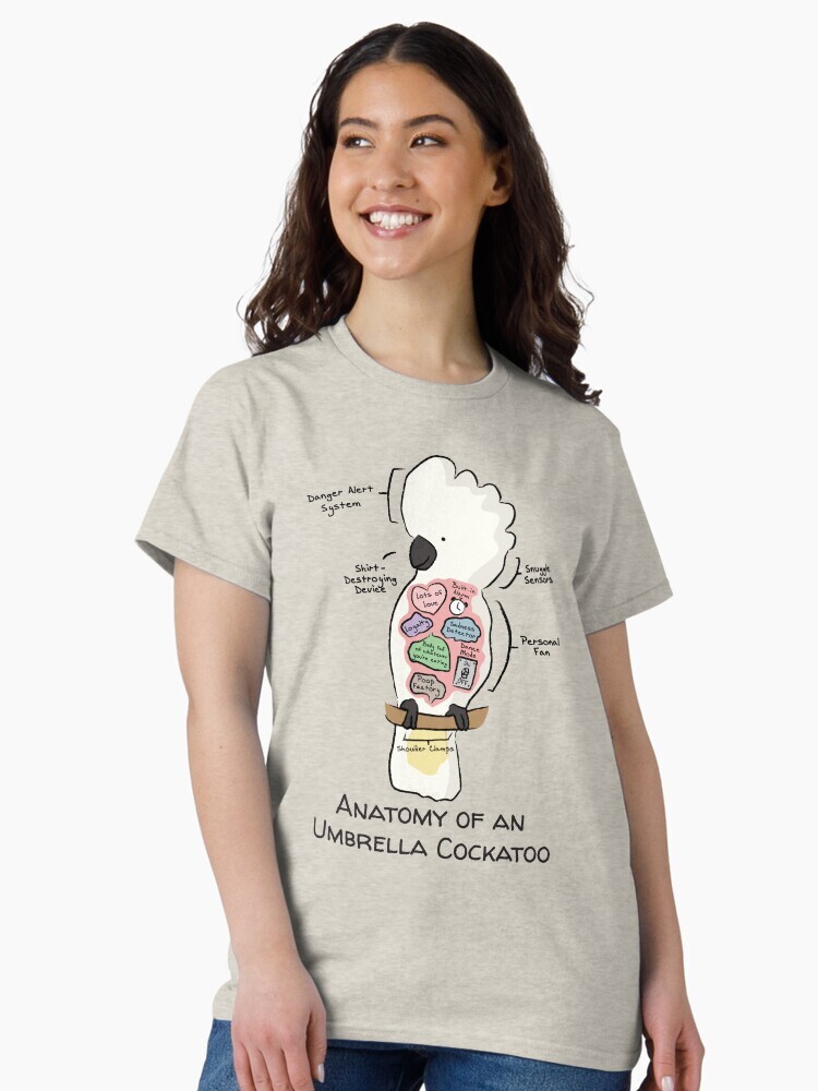 A brunette woman wearing a t-shirt  featuring a cartoon illustration titled “Anatomy of an Umbrella Cockatoo” showing a cute Umbrella Cockatoo bird with funny labeled anatomy sections.