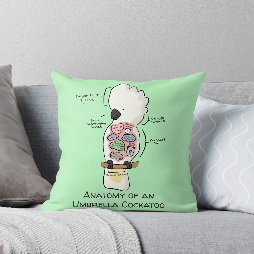 Decorative throw pillow on a couch featuring a cartoon illustration titled “Anatomy of an Umbrella Cockatoo” showing a cute Umbrella Cockatoo with funny labeled anatomy sections.
