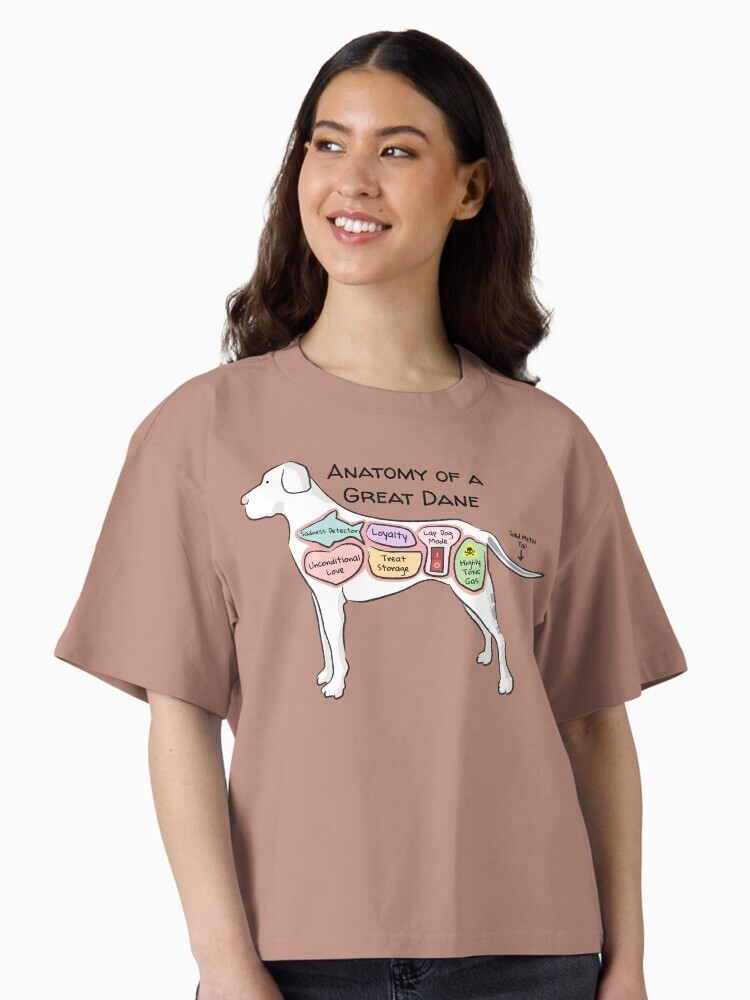A brunette woman wearing a t-shirt  featuring a cartoon illustration titled “Anatomy of a Great Dane” showing a cute Great Dane dog with funny labeled anatomy sections.