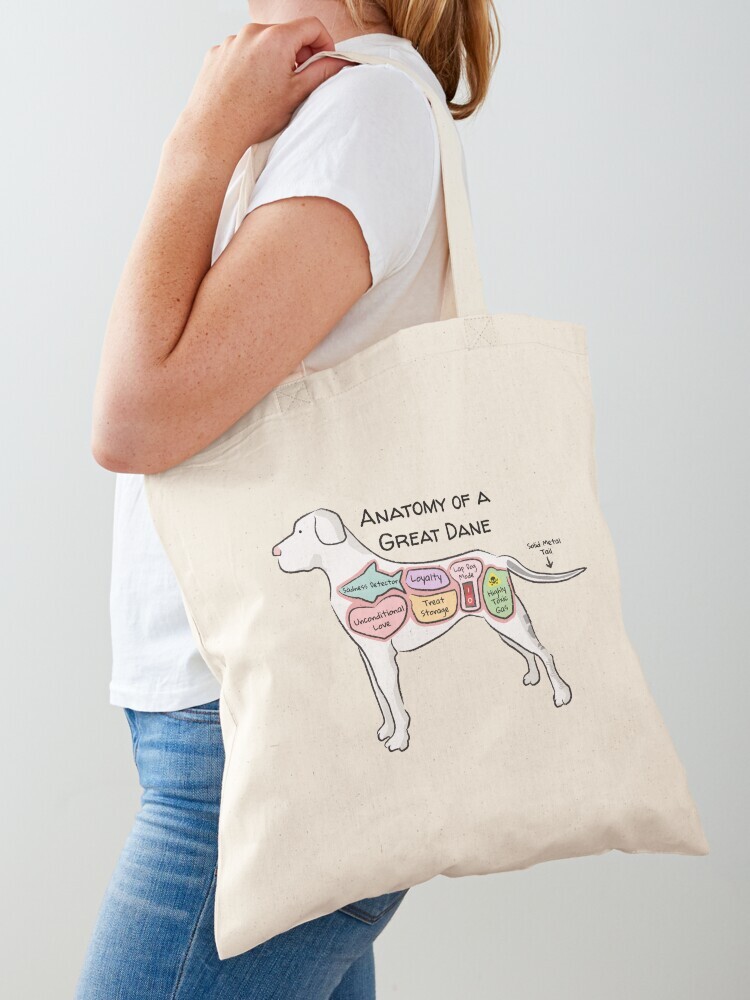 A woman wearing a white short sleeve t-shirt with a cotton tote bag on her shoulder. The tote bag features a cartoon illustration titled “Anatomy of a Great Dane” showing a cute Great Dane dog with funny labeled anatomy sections.