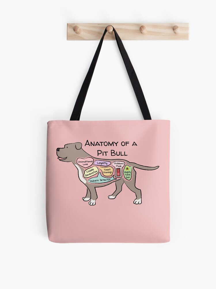 A pink-colored tote bag hanging off a wooden hook. On the tote bag features a cartoon illustration titled “Anatomy of a Pit Bull” showing a cute Pit Bull dog with funny labeled anatomy sections.