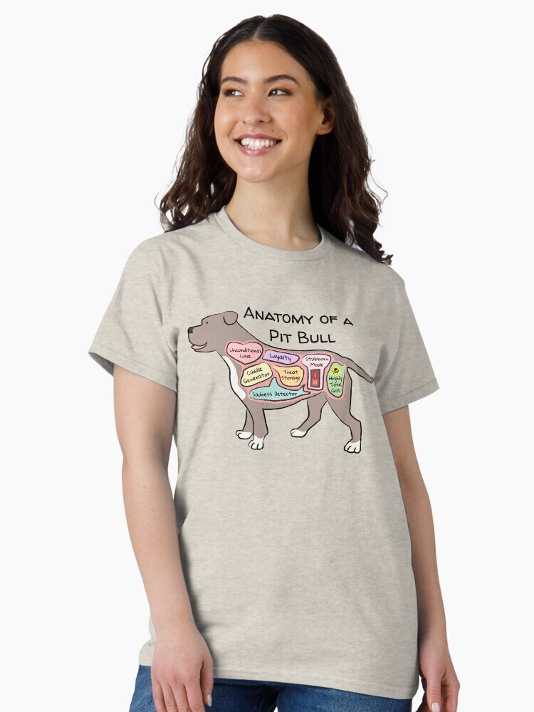 A brunette woman wearing an oversized t-shirt  featuring a cartoon illustration titled “Anatomy of a Pit Bull” showing a cute Pit Bull dog with funny labeled anatomy sections.