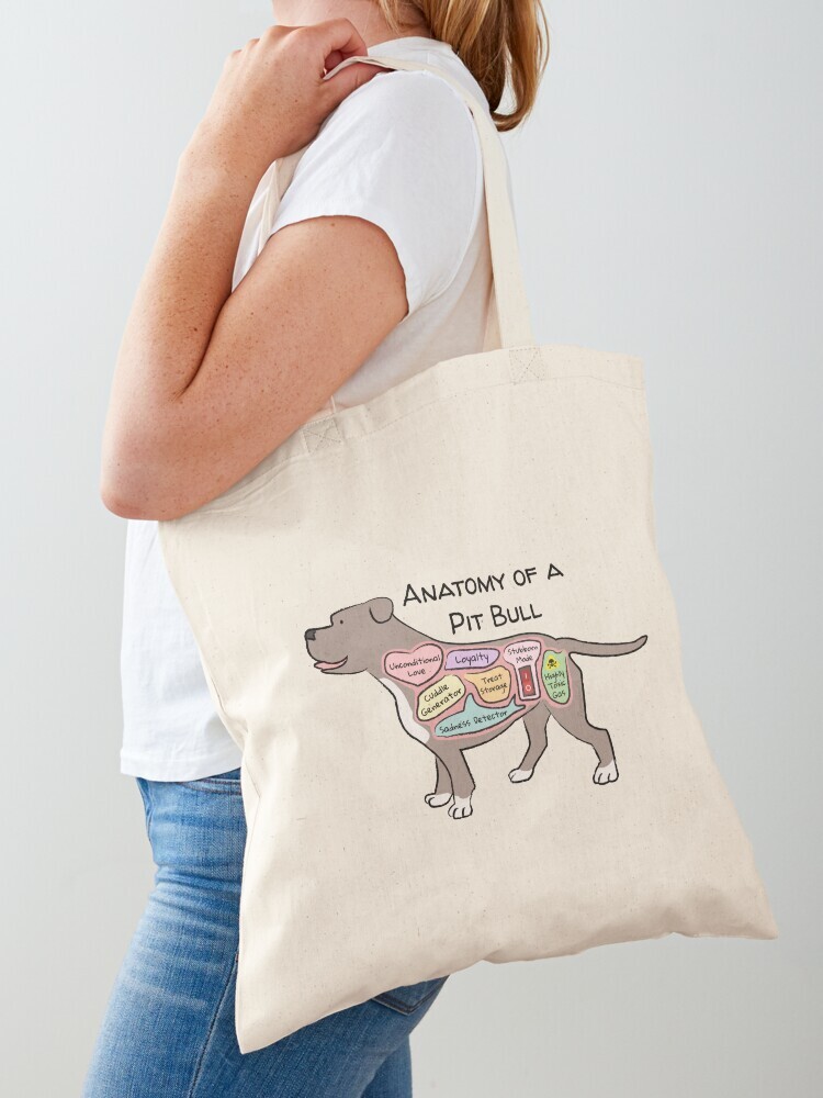 A woman wearing a white short sleeve t-shirt with a cotton tote bag on her shoulder. The tote bag features a cartoon illustration titled “Anatomy of a Pit Bull” showing a cute Pit Bull dog with funny labeled anatomy sections.