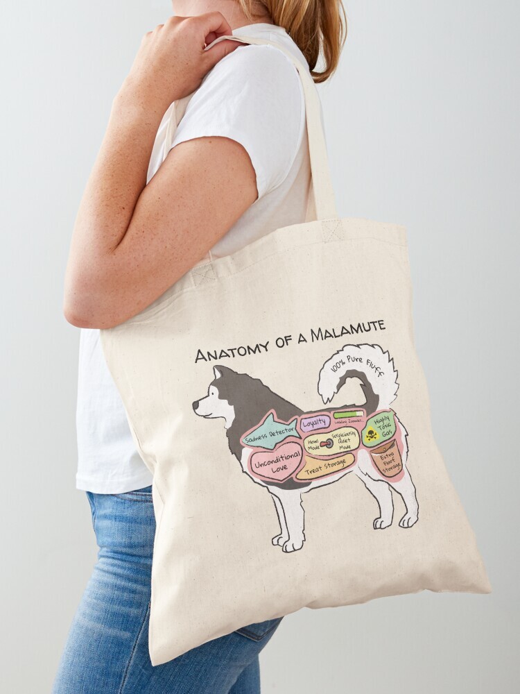 A woman wearing a white short sleeve t-shirt with a cotton tote bag on her shoulder. The tote bag features a cartoon illustration titled “Anatomy of a Malamute” showing a cute Malamute dog with funny labeled anatomy sections.