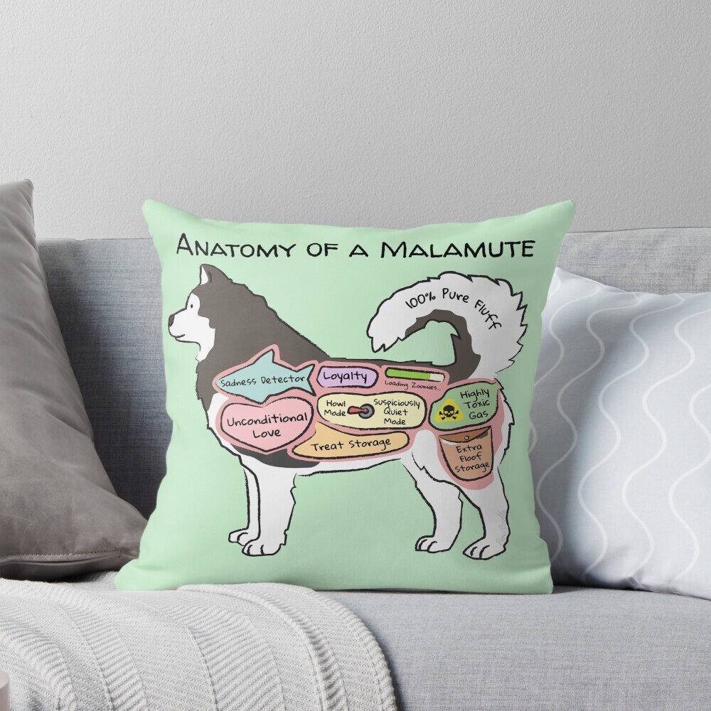 Decorative throw pillow on a couch featuring a cartoon illustration titled “Anatomy of a Malamute” showing a cute Malamute dog with funny labeled anatomy sections.
