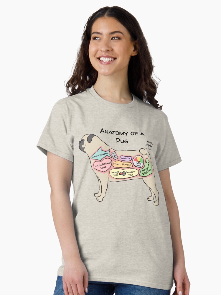 A brunette woman wearing a beige t-shirt  featuring a cartoon illustration titled “Anatomy of a Pug” showing a cute Pug dog with funny labeled anatomy sections.