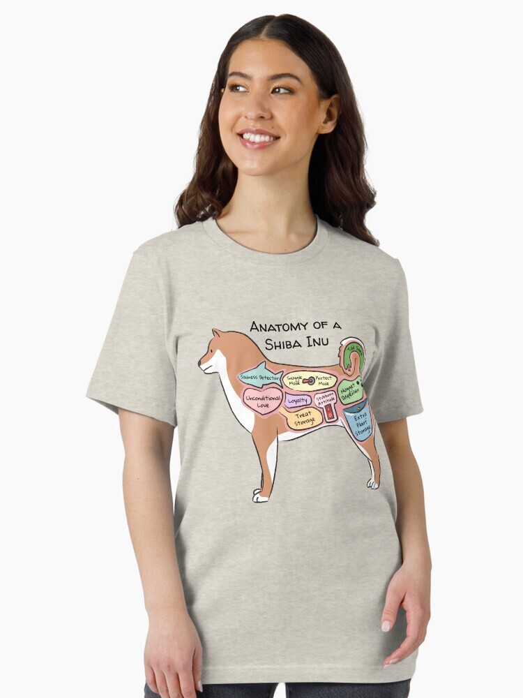 A brunette woman wearing a t-shirt  featuring a cartoon illustration titled “Anatomy of a Shiba Inu” showing a cute Shiba Inu dog with funny labeled anatomy sections.