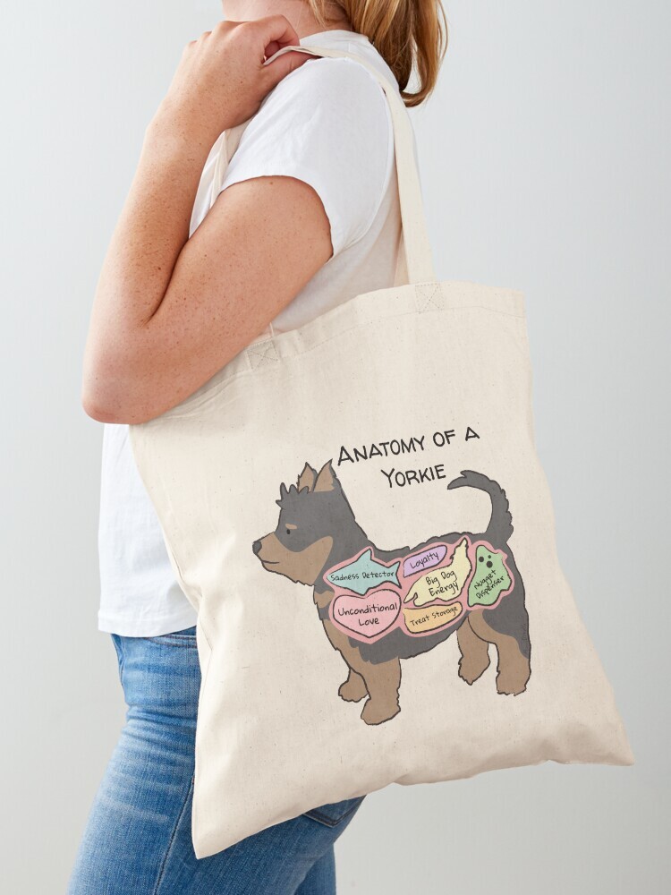 A woman wearing a white short sleeve t-shirt with a cotton tote bag on her shoulder. The tote bag features a cartoon illustration titled “Anatomy of a Yorkie” showing a cute Yorkhire Terrier dog with funny labeled anatomy sections.